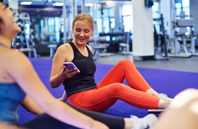 Woman in a gym holding a smartphone, with gym equipment in the background.
